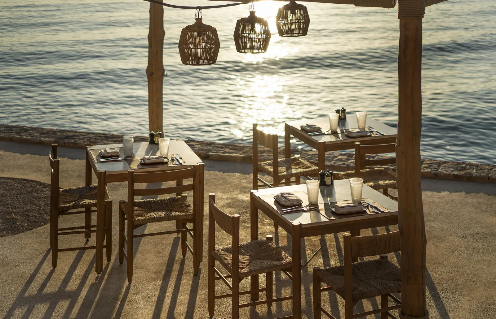 The wooden tables of Taverna 37 on the shore at sunset, under a pergola hung with wicker lamps, a step from the water.