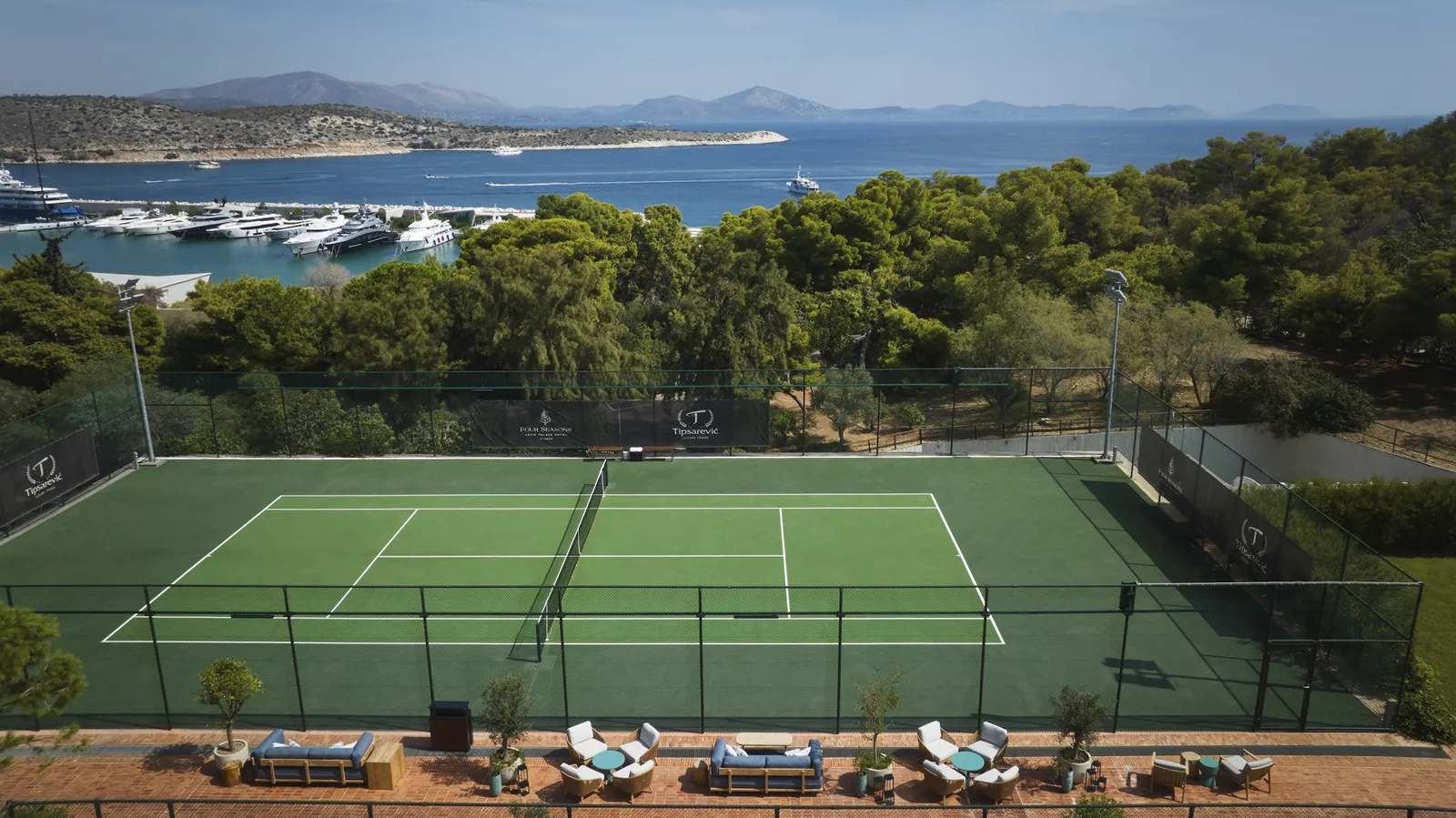 A green tennis court among the pines, with a row of armchairs at the side and, behind the trees, the marina and the gulf.