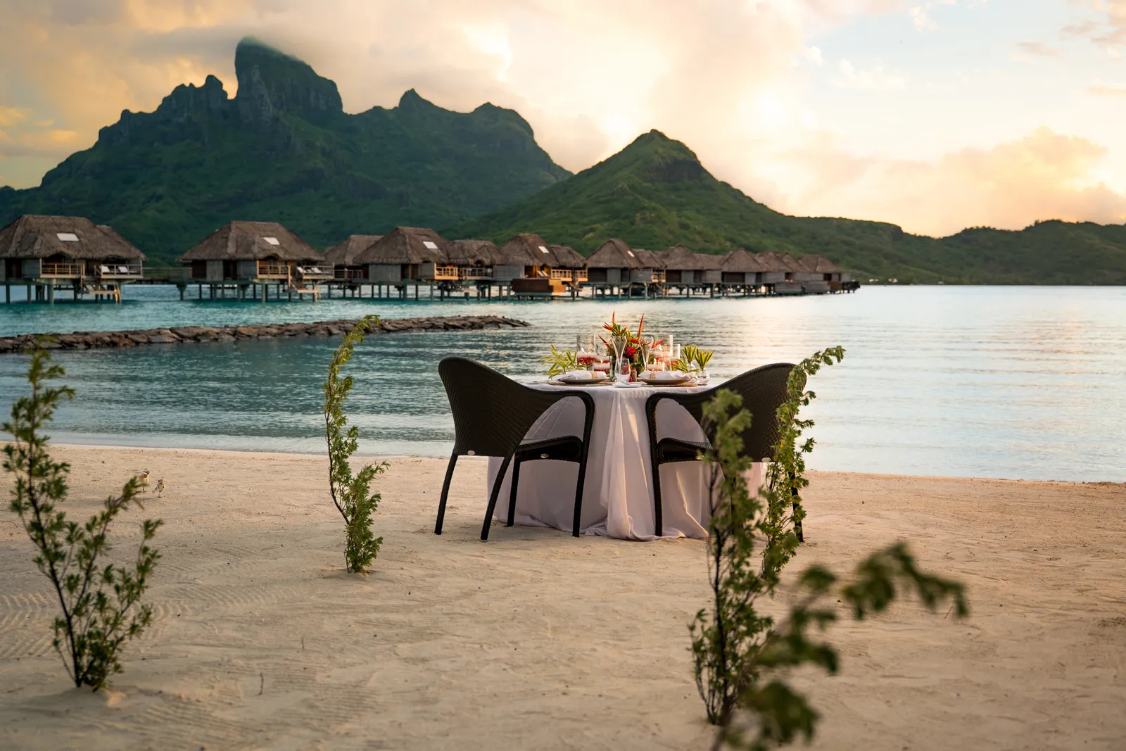 A table for 2 laid on the sand at sunset, the bungalows and Otemanu behind