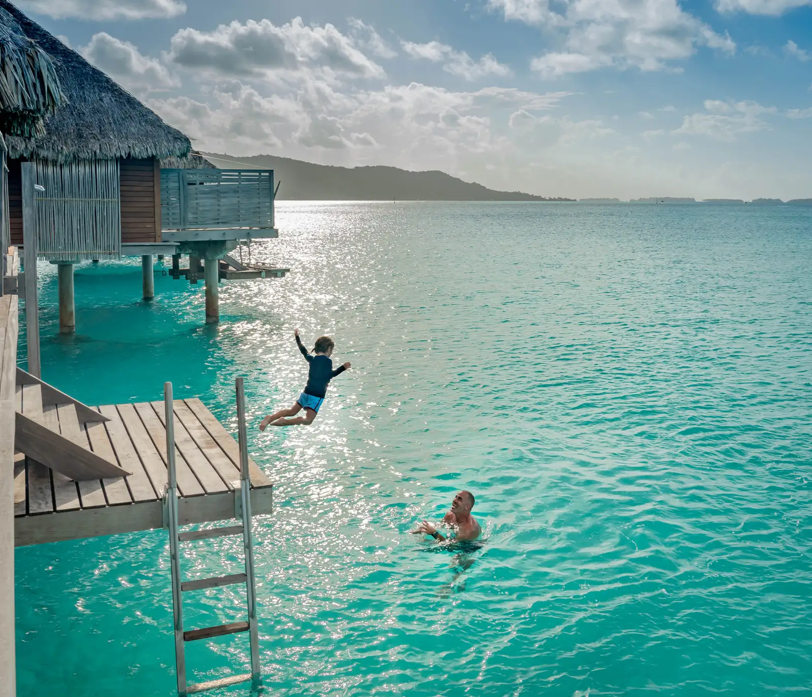 A child leaps from a bungalow deck into the lagoon, an adult waiting in the water