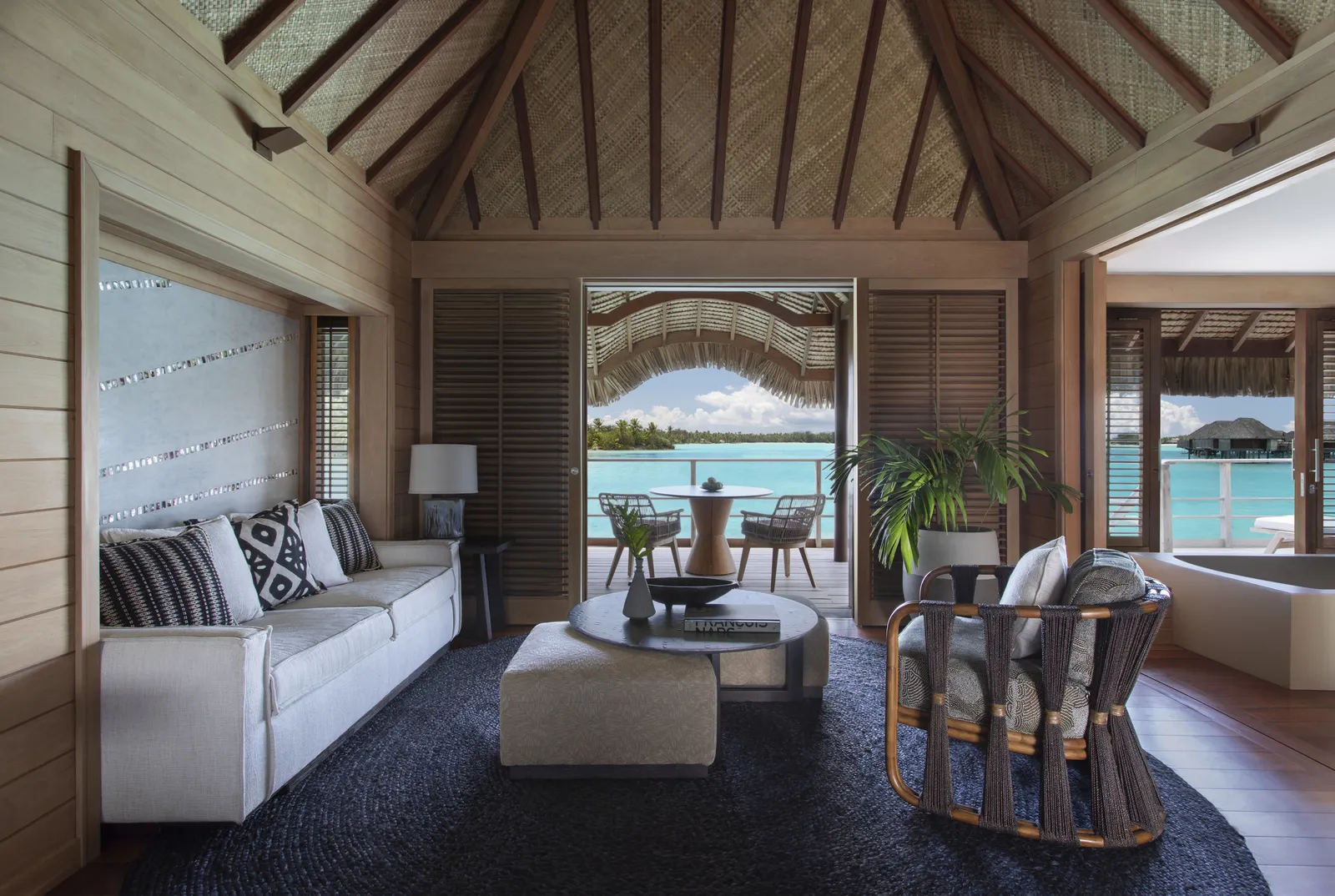 An overwater suite’s sitting room: a white sofa and the lagoon beyond the glass doors