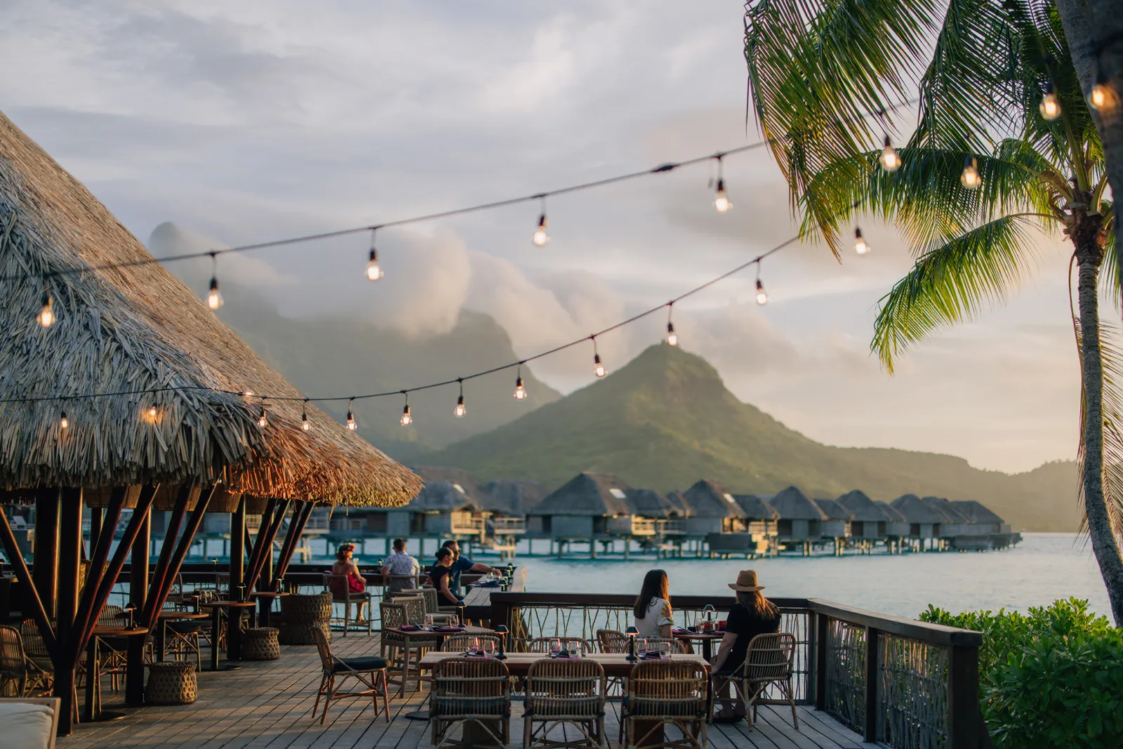 A terrace under string lights at dusk, overwater bungalows and the island’s peaks behind