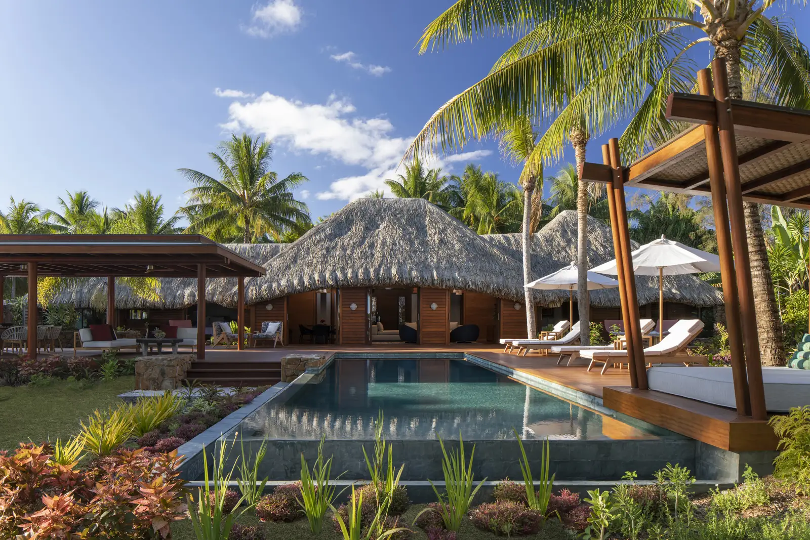 A villa pool in its garden, with loungers and thatched pavilions