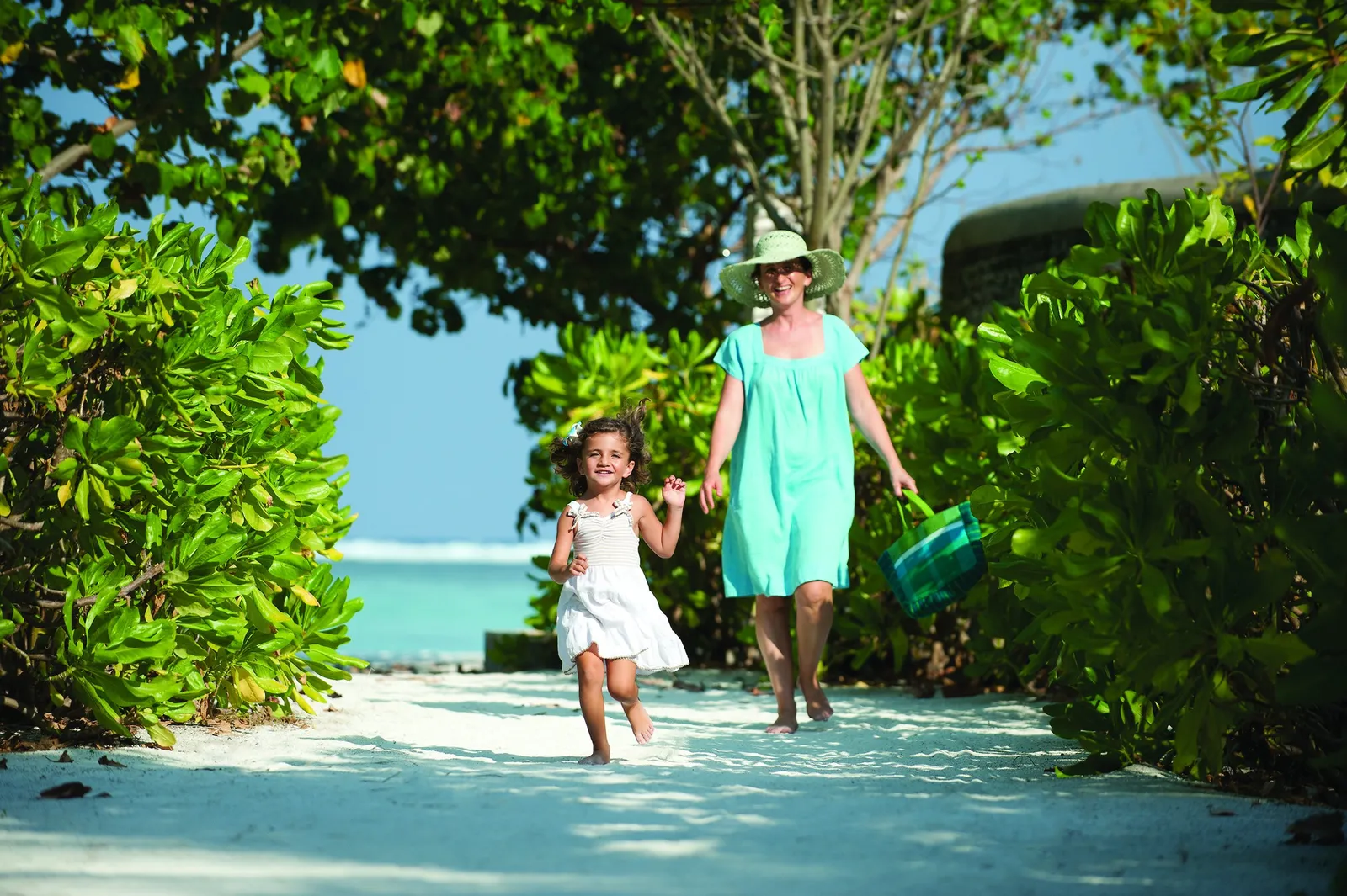 A mother and a little girl running down a sandy path through the greenery to the beach
