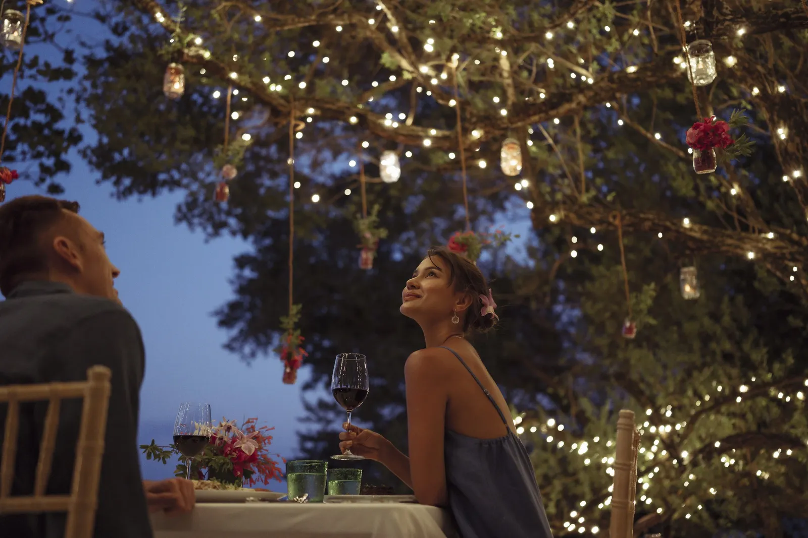 A couple at dinner under a tree hung with fairy lights and jars of flowers