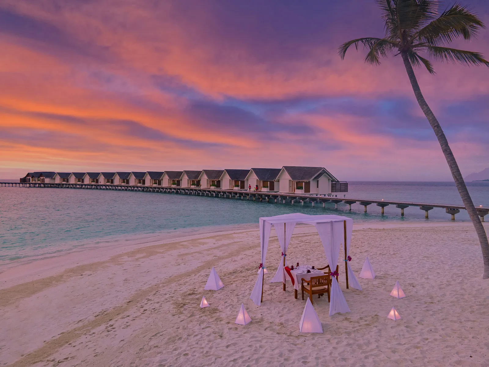 A table for 2 on the beach at sunset among lanterns, the villa jetty beyond
