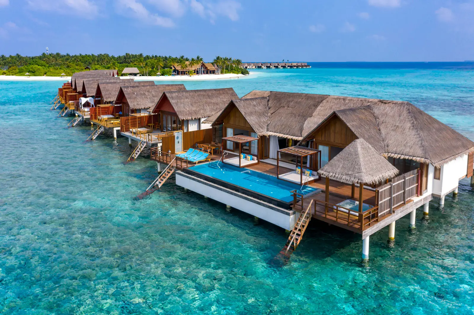The overwater residences from the air, pools on their decks and the island’s beach behind