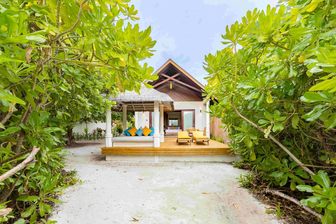 A Beach Villa seen from the sandy path between 2 bushes, its daybed under a thatched roof