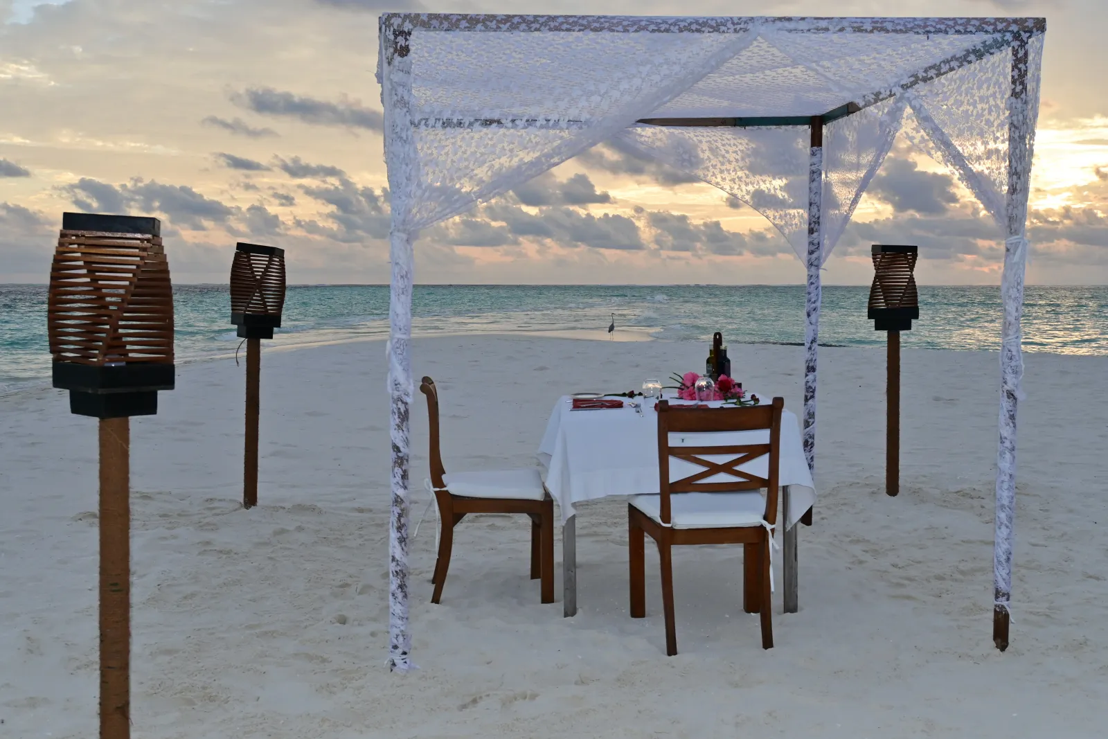 A table for 2 on the beach under a lace canopy, among the torches, at sunset.
