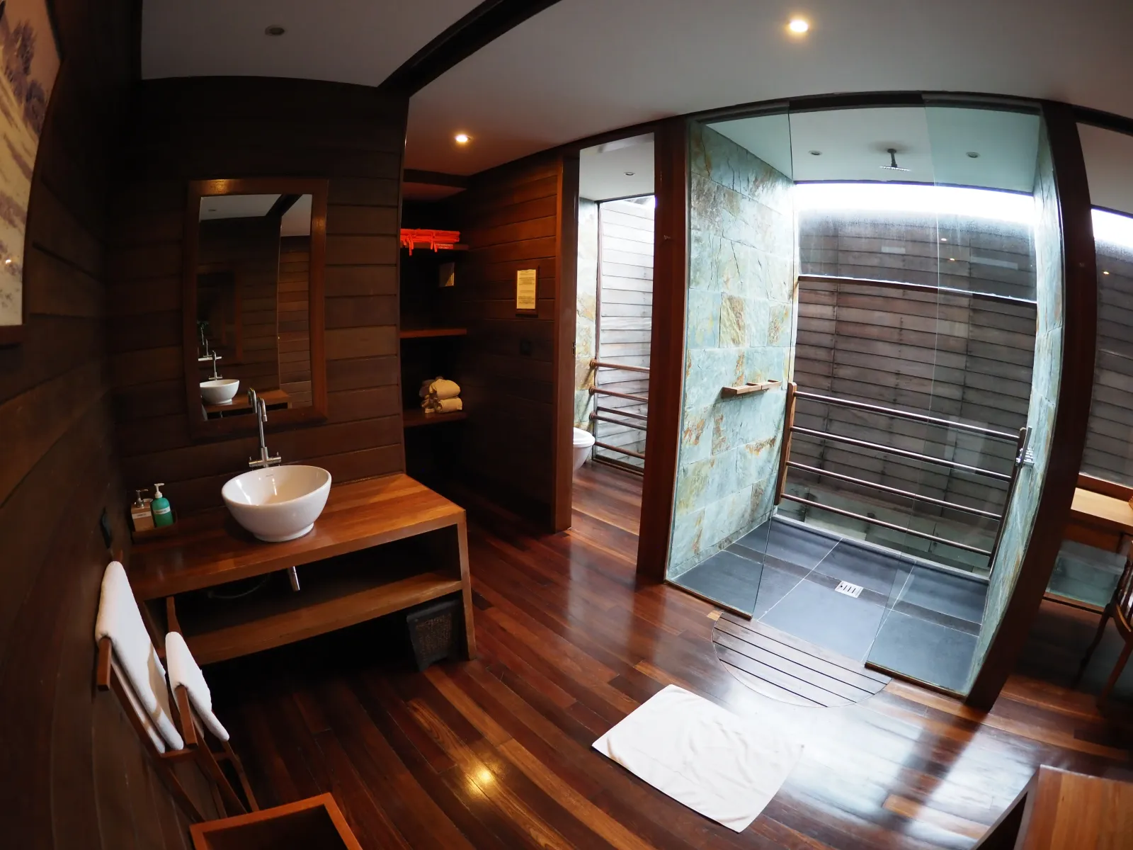 An overwater villa bathroom in dark wood, with a vessel basin and the shower behind glass.