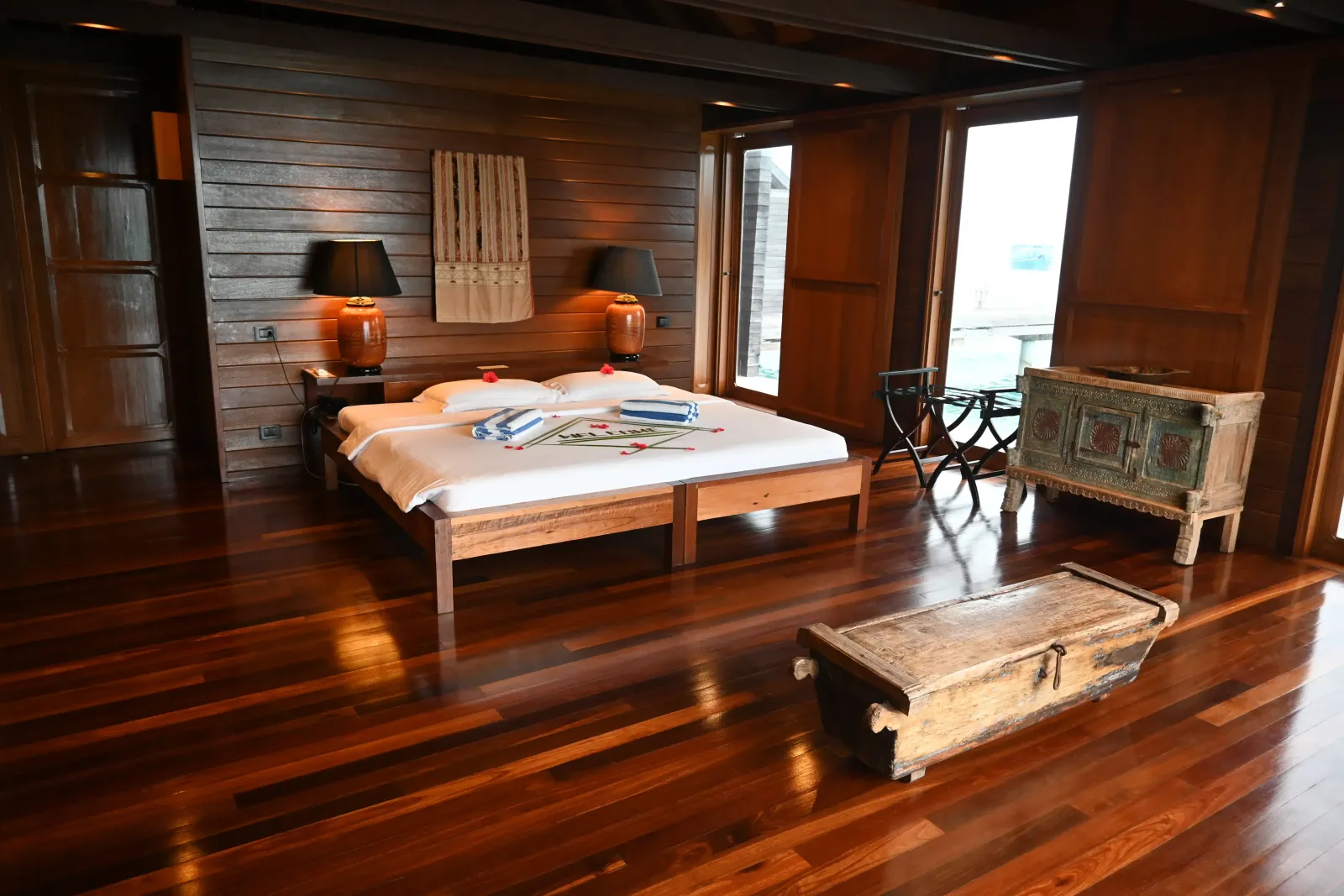 Inside an Overwater Villa Deluxe: the bed, the polished wooden floor, a carved chest and the lamps.