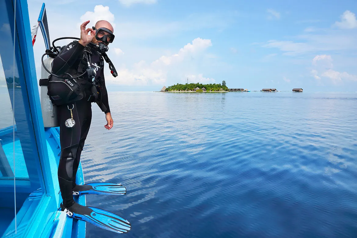 A diver in a wetsuit steps off the dive centre’s boat into the lagoon, with an island on the horizon.