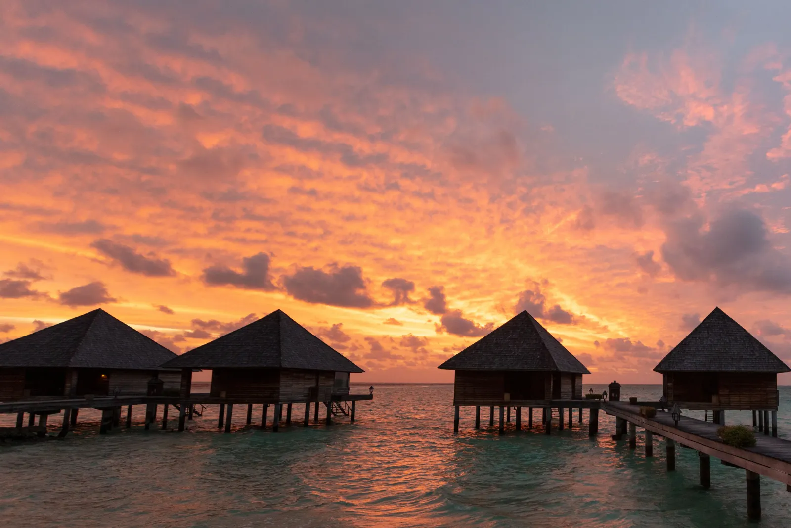 The overwater villas with their pointed roofs, in silhouette, under an orange sky after sunset.