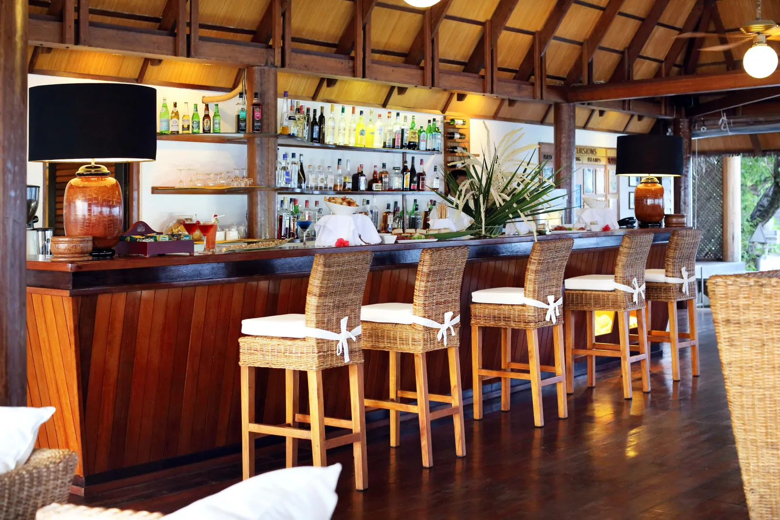 The dark wood counter of the Kandu Bar, with wicker stools and bottles on the shelves.
