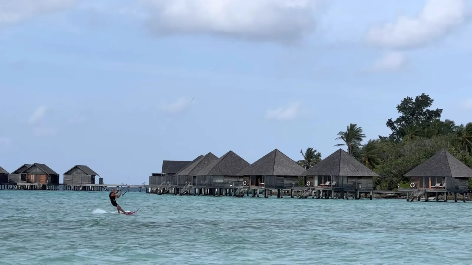 A kitesurfer in the lagoon in front of the row of overwater villas and the palms.
