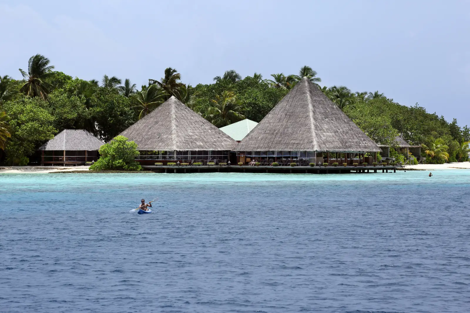 The great pyramid roofs of the pavilions on the shore, among the palms, seen from the lagoon with a canoe on the water.
