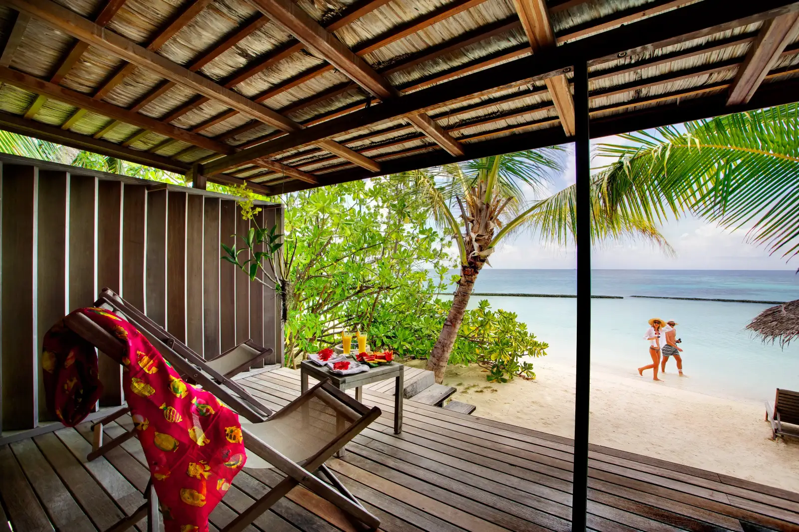A Beach Villa’s covered veranda, with a deckchair and a small table, and ahead the palms, the beach and a couple walking.