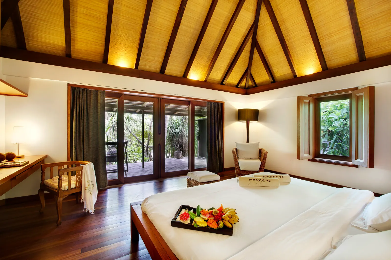 A Garden Villa: the bed with a fruit plate, the pitched timber ceiling and glass doors open on the greenery.