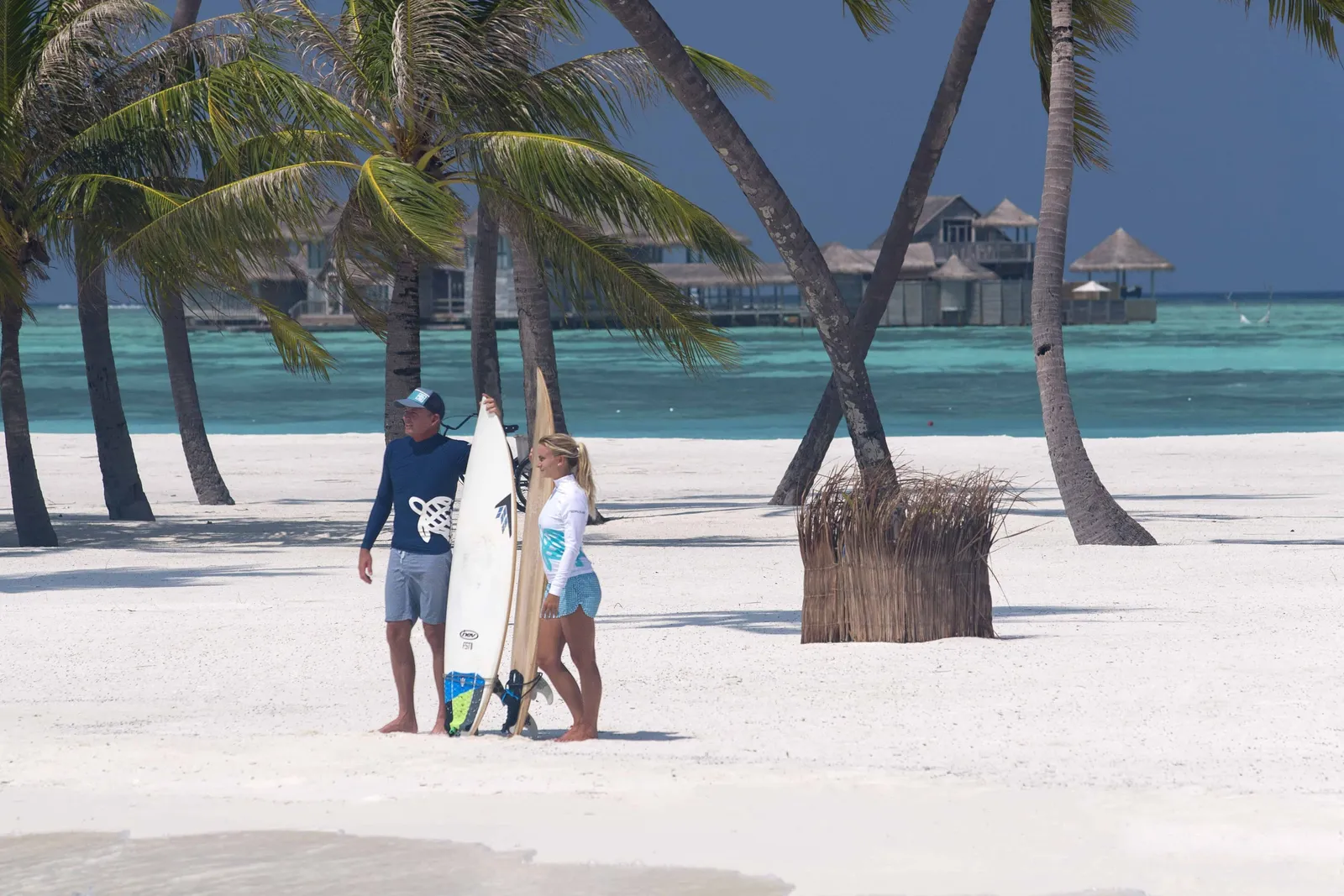 2 surfers on the beach with their boards, the overwater villas behind