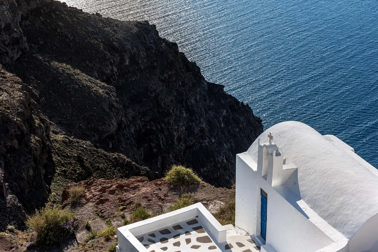 A white chapel with a blue door and a cross on its bell gable, below the hotel, against the dark rock face and the sea.