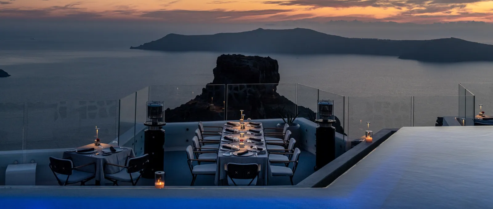 A long table laid by candlelight beside the pool after sunset, with the dark outline of Skaros.