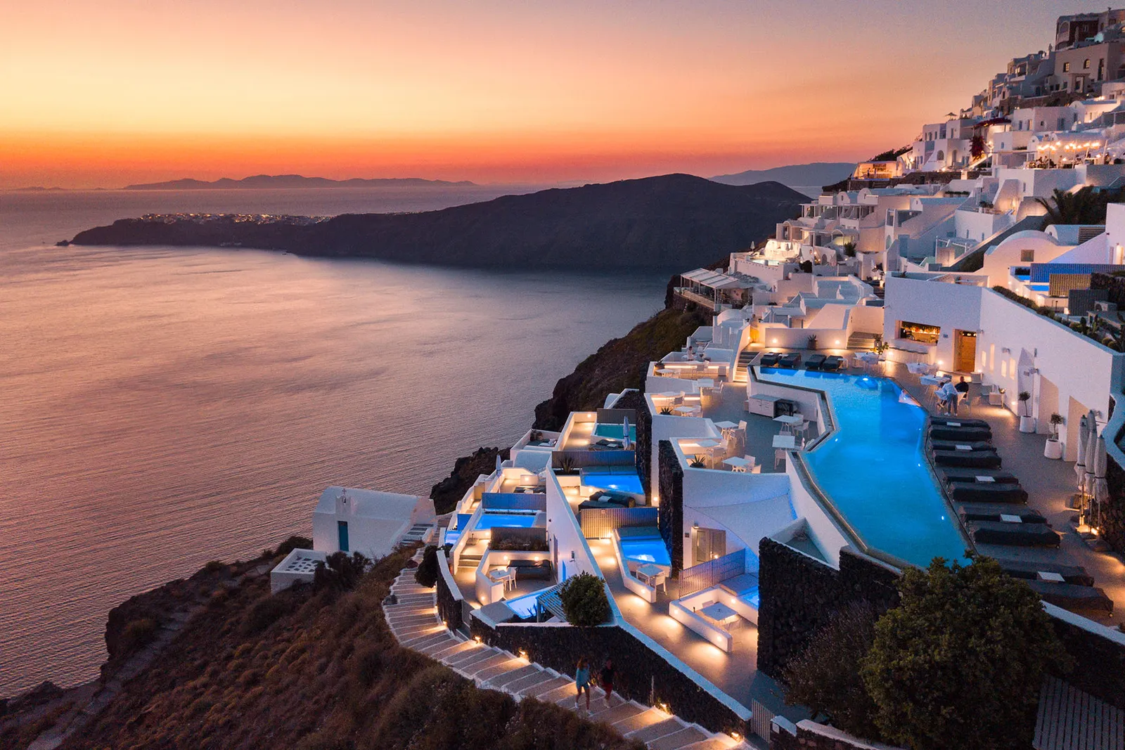 The hotel from above at nightfall: lit terraces stepping down the cliff, the blue pool and an orange sky over the sea.