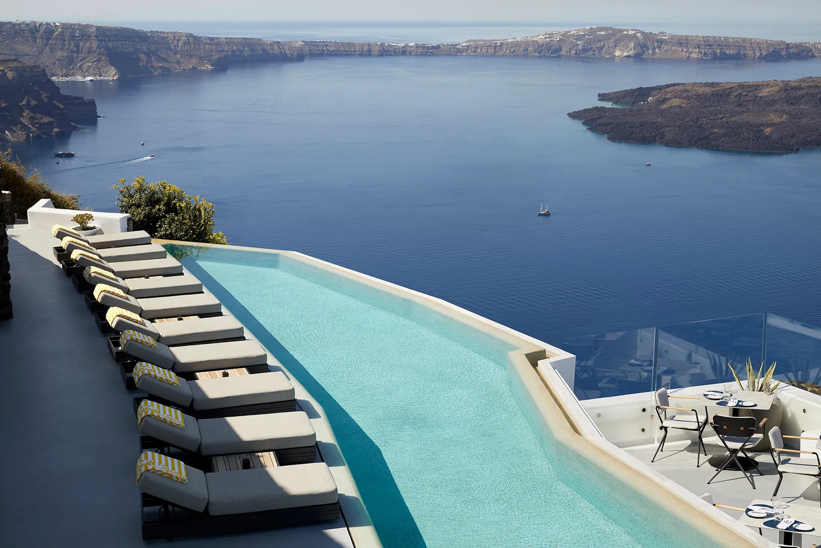 The row of loungers along the infinity pool, seen from above, with the dark blue caldera and its cliffs.