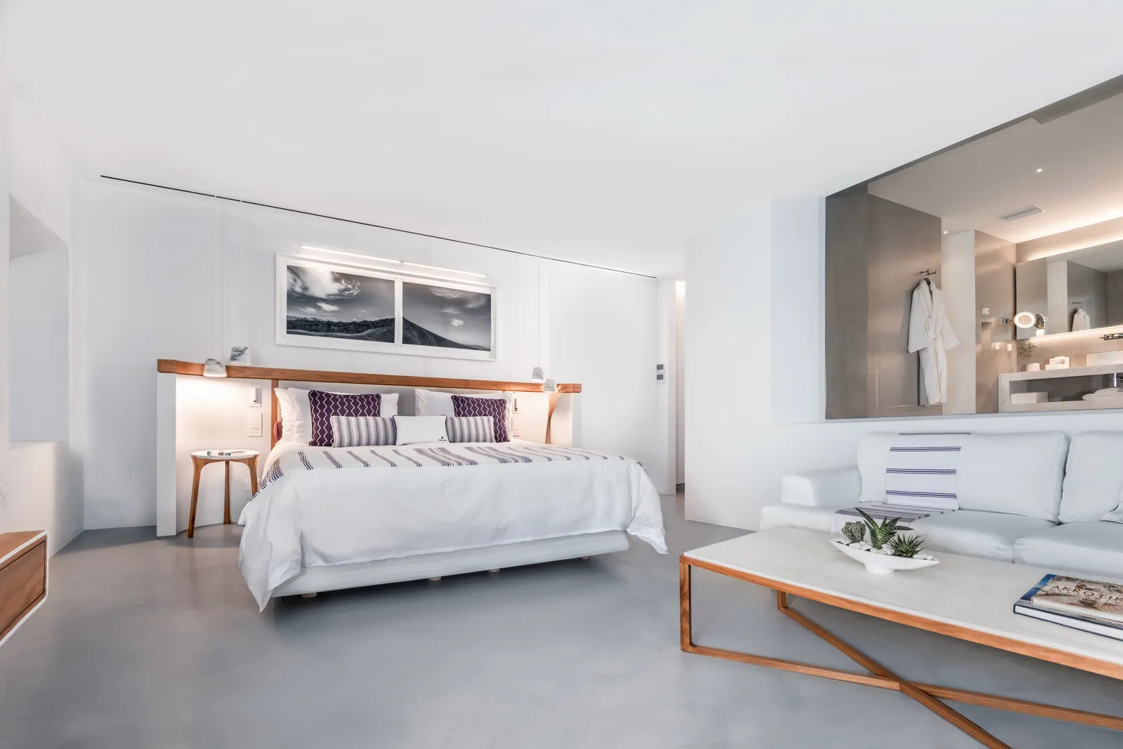 A Grace Suite’s bedroom: the bed with purple cushions, the white sofa, a low table and the open bathroom behind a grey wall.