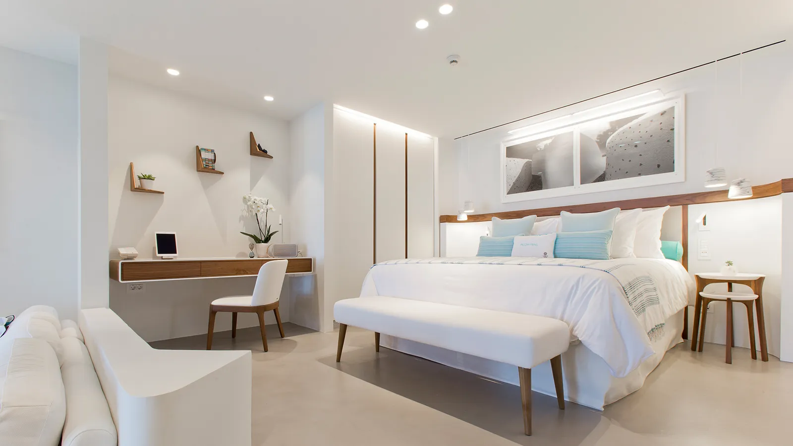 The VIP Suite’s bedroom: the white bed with pale blue cushions, a bench at its foot, the wooden desk and a white armchair.