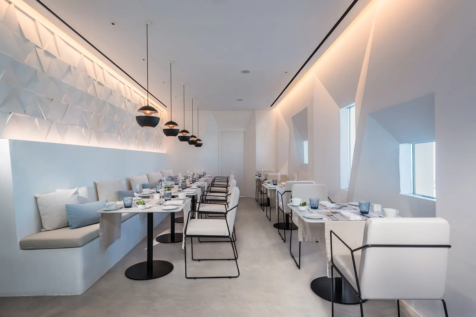 Varoulko Santorini’s dining room: tables laid in a row, white chairs, black pendant lamps and a white wall in relief.