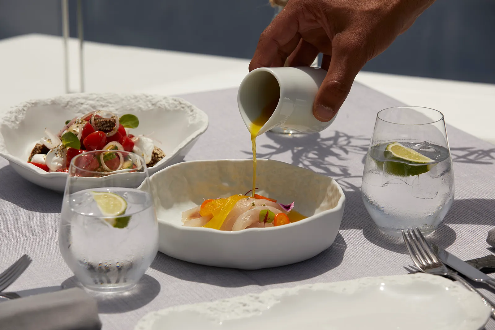 A hand pours olive oil over a plate of raw fish, beside a tomato salad and 2 glasses of water with lime.