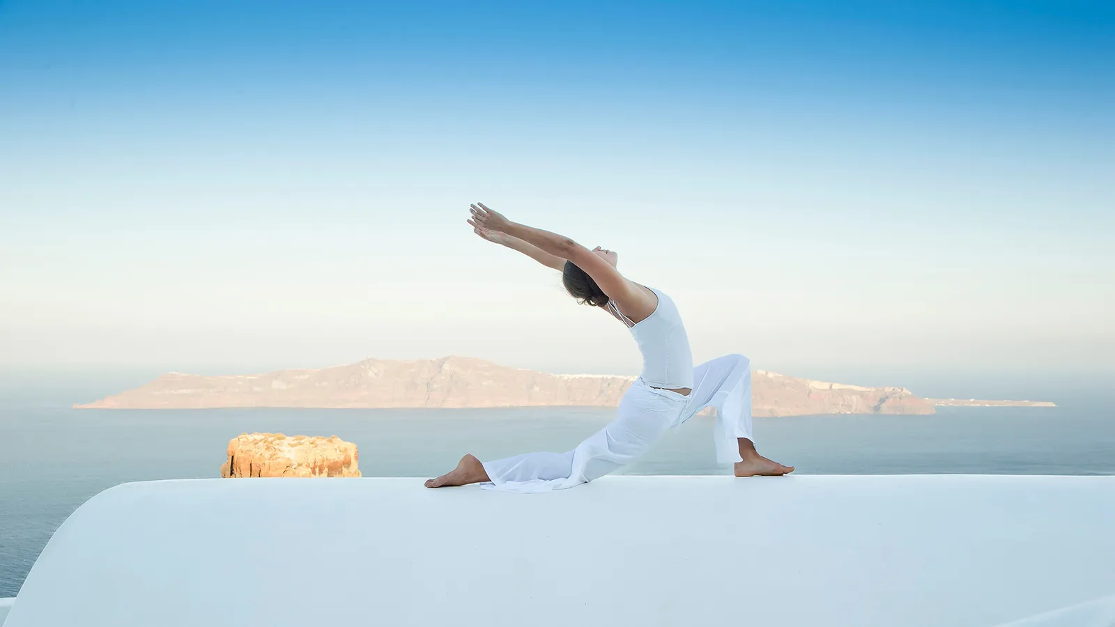 A figure in white holding a yoga pose on a white terrace, the sea and an island on the horizon.
