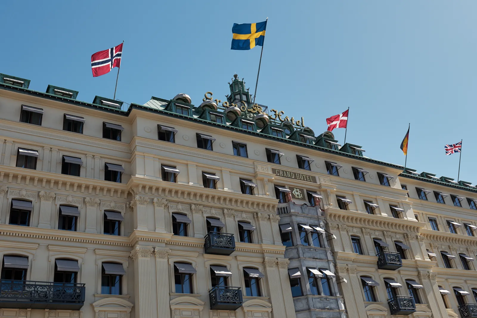 The hotel’s façade from below on a sunny day: dark awnings at the windows, iron balconies and the Scandinavian flags on the roof.