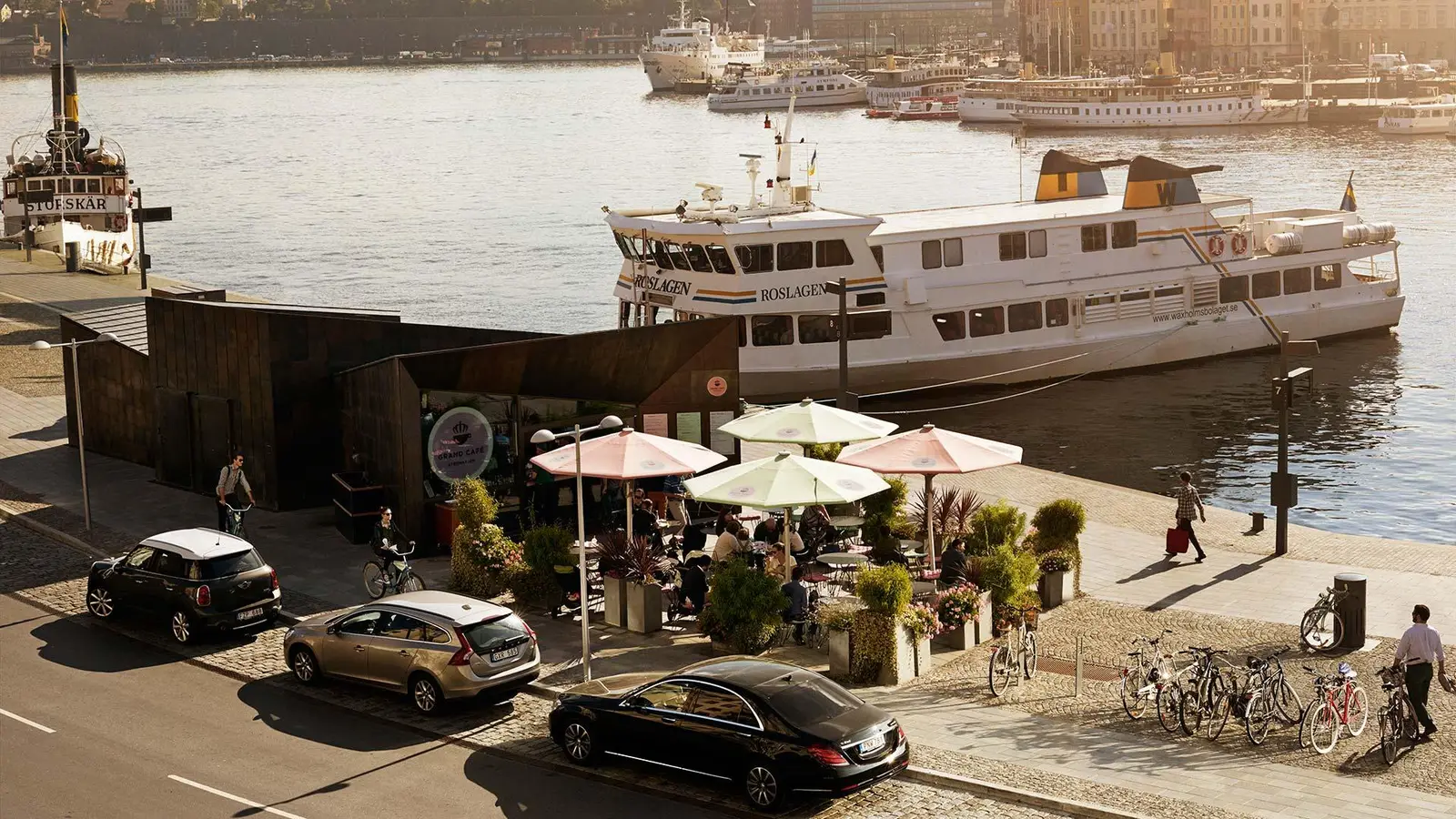 The Grand Café on the quay before the hotel: pale parasols among the plants, a white archipelago boat moored and low light on the water.