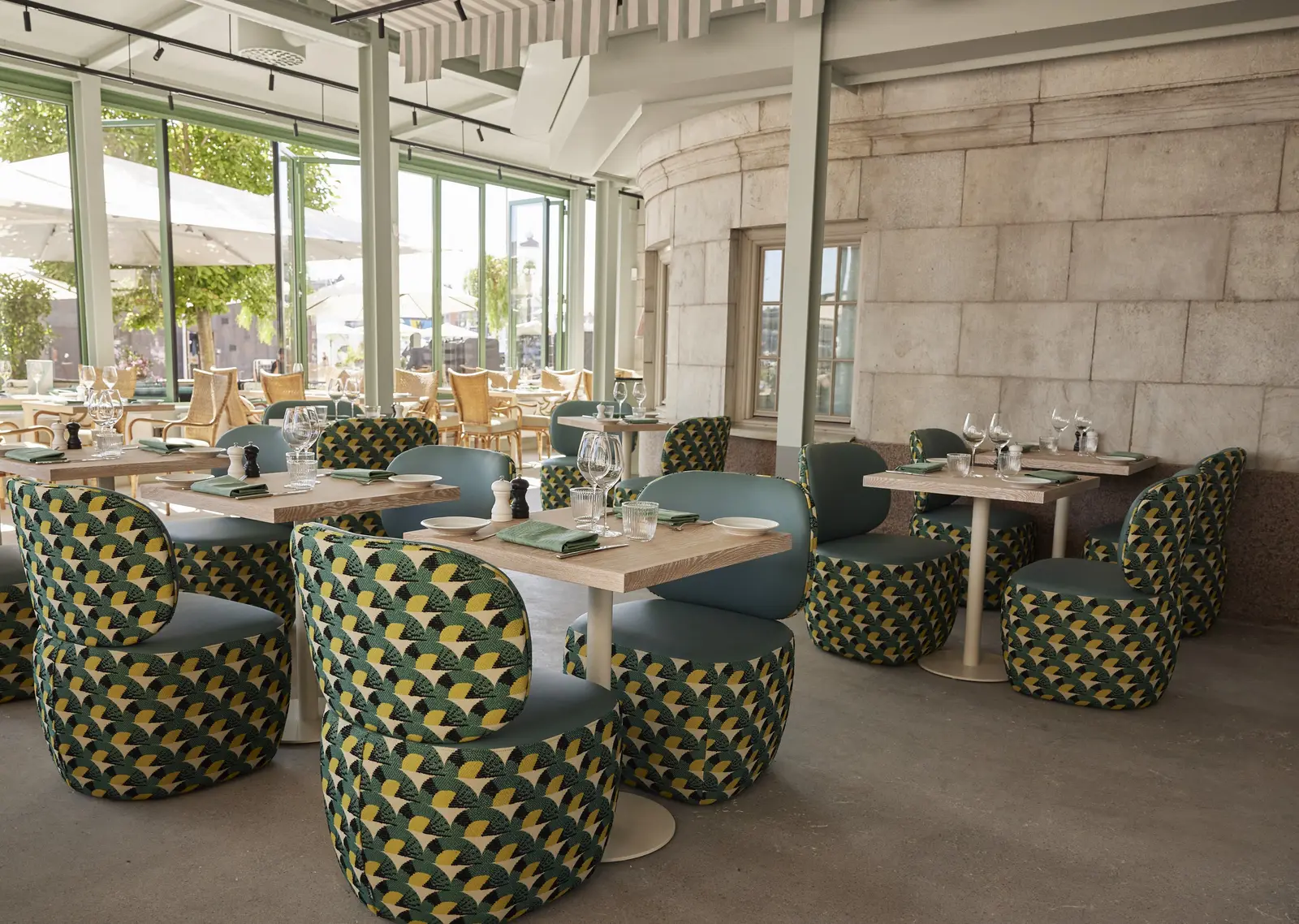 The Grand Soleil dining room: tub chairs in green and yellow fans around pale wooden tables, under the glass veranda, the terrace beyond.