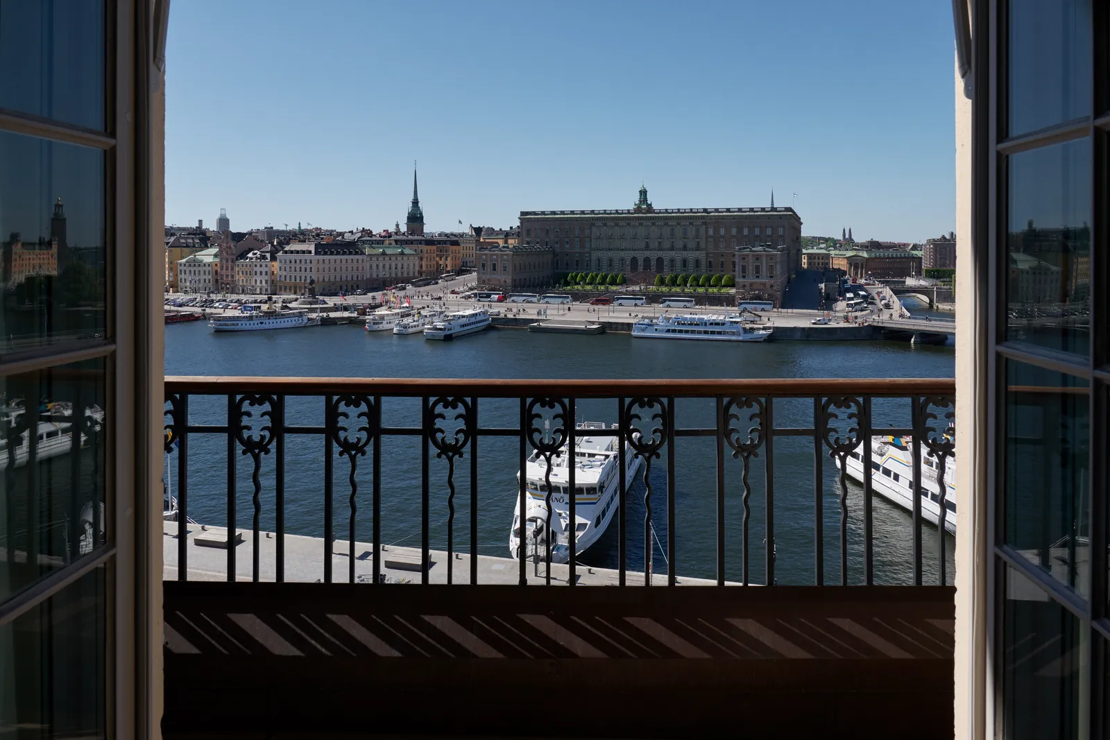 Through open French windows onto a wrought-iron balcony, the Royal Palace and the roofs of Gamla Stan across the water, white boats moored at the quay.