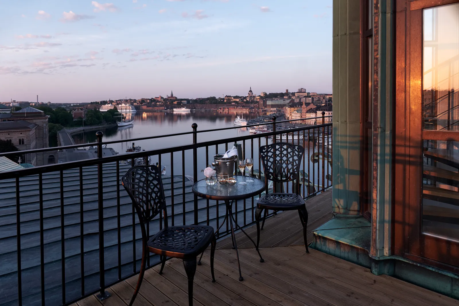 A small table with 2 iron chairs and a Champagne bucket on a rooftop terrace at dusk, the harbour and the city lights on the water.