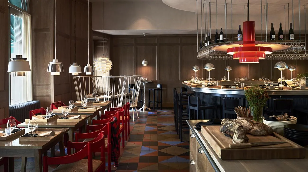 Matbaren’s dining room: wooden tables with red chairs, the curved counter set with plates, white pendant lamps and one red lamp above the bar.