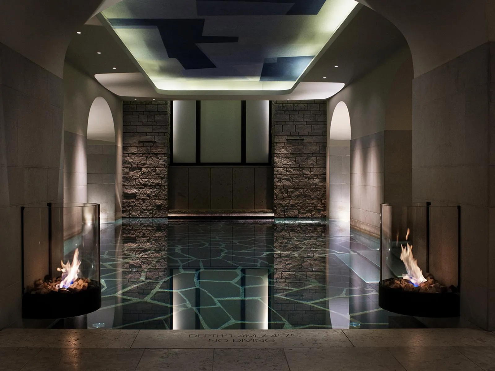 The spa pool: a dark basin between walls of split stone, 2 lit fires behind glass and a ceiling lit in panels.