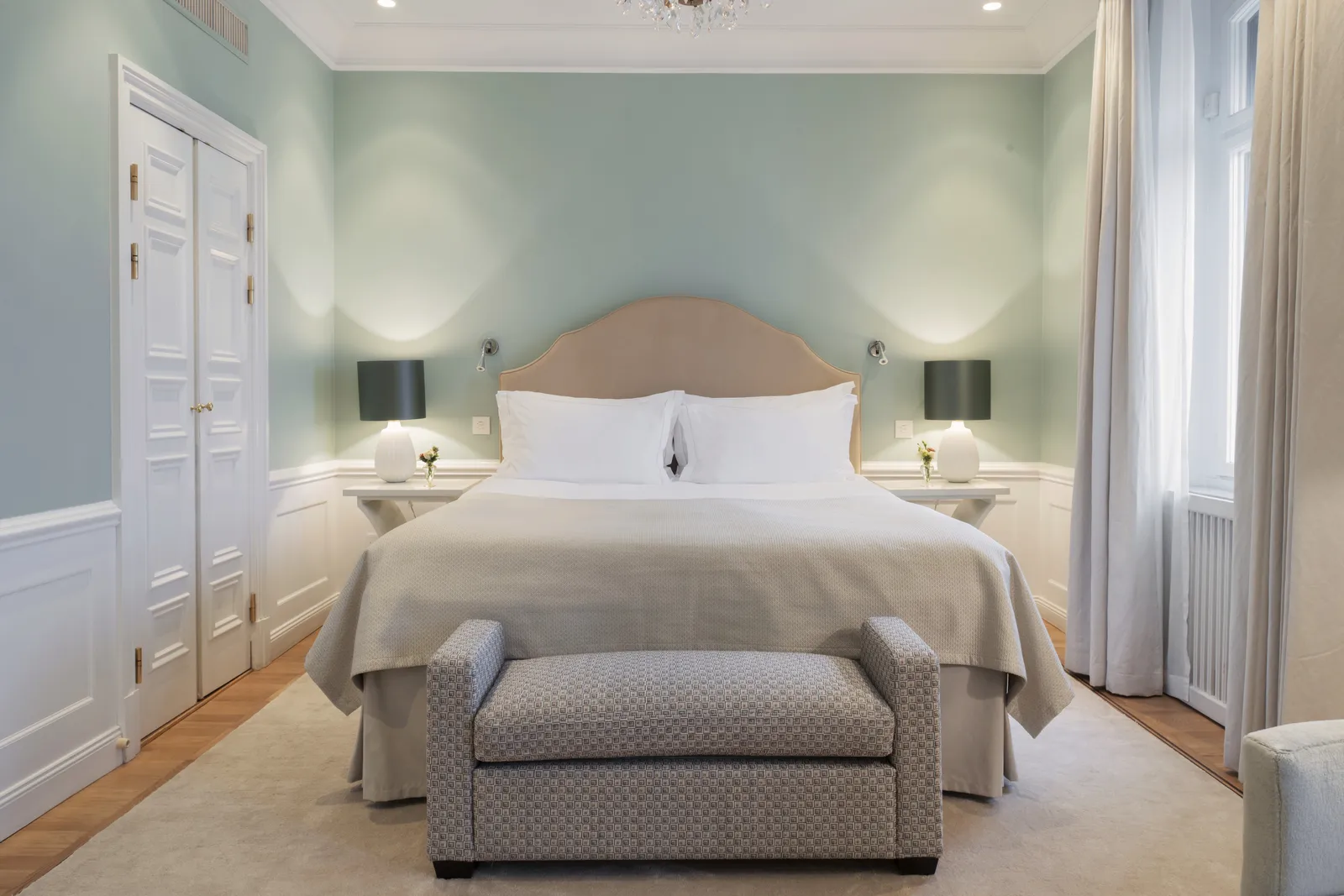 A water-view Deluxe Room: the king bed with a pale headboard, pale-green walls above white panelling and a bench at the foot of the bed.