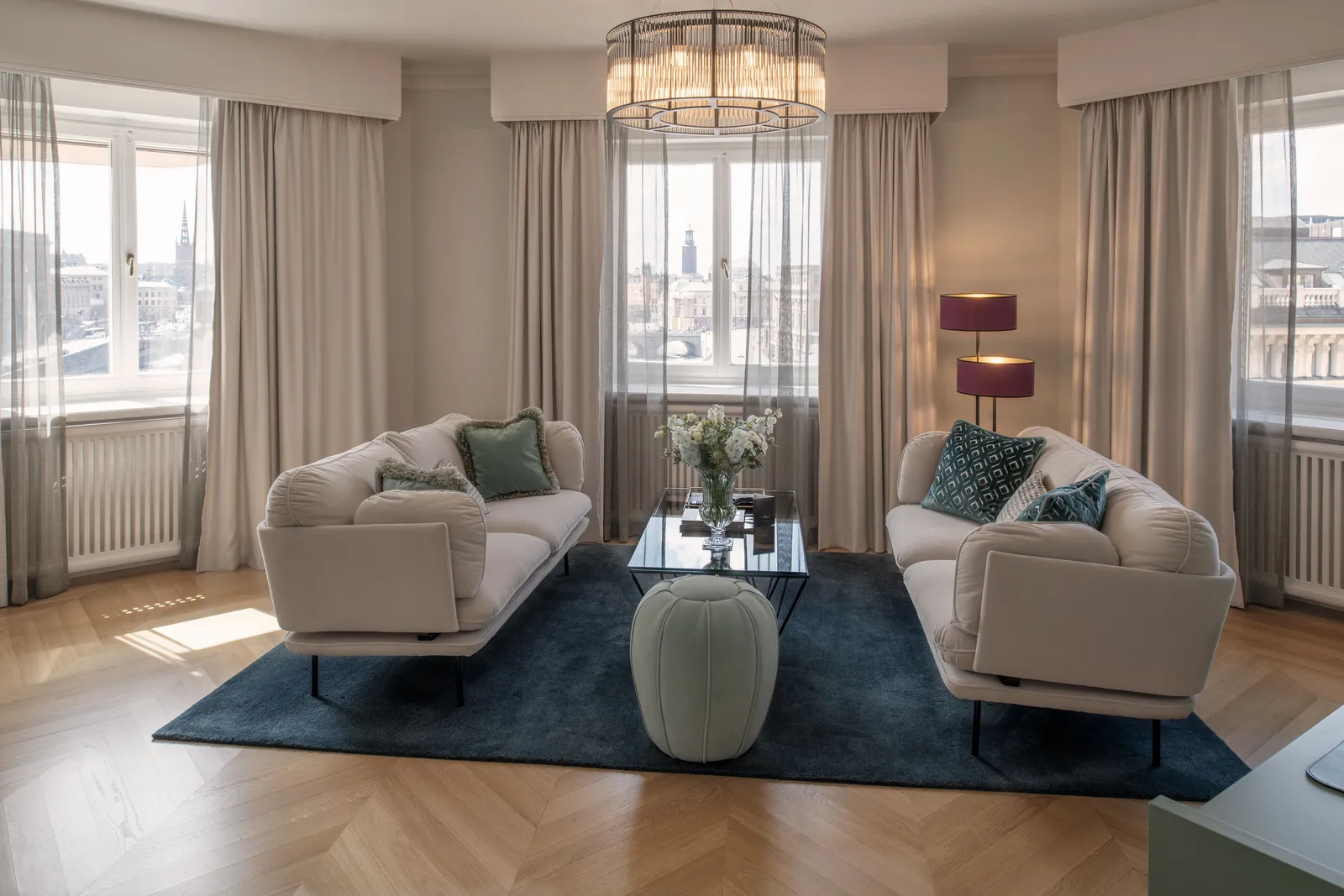 An Executive Suite sitting room: 2 pale sofas on a blue rug, a green pouffe and 3 windows with the city beyond the curtains.