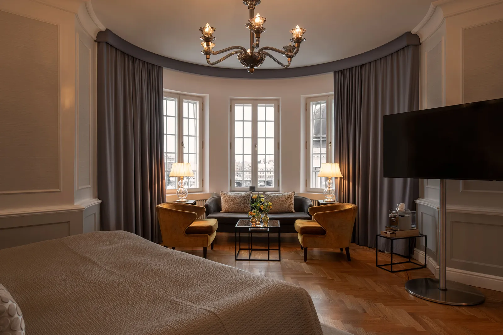 A Junior Suite: the sitting area in a bay of 3 windows, 2 mustard armchairs around a low table and, in the foreground, the bed.