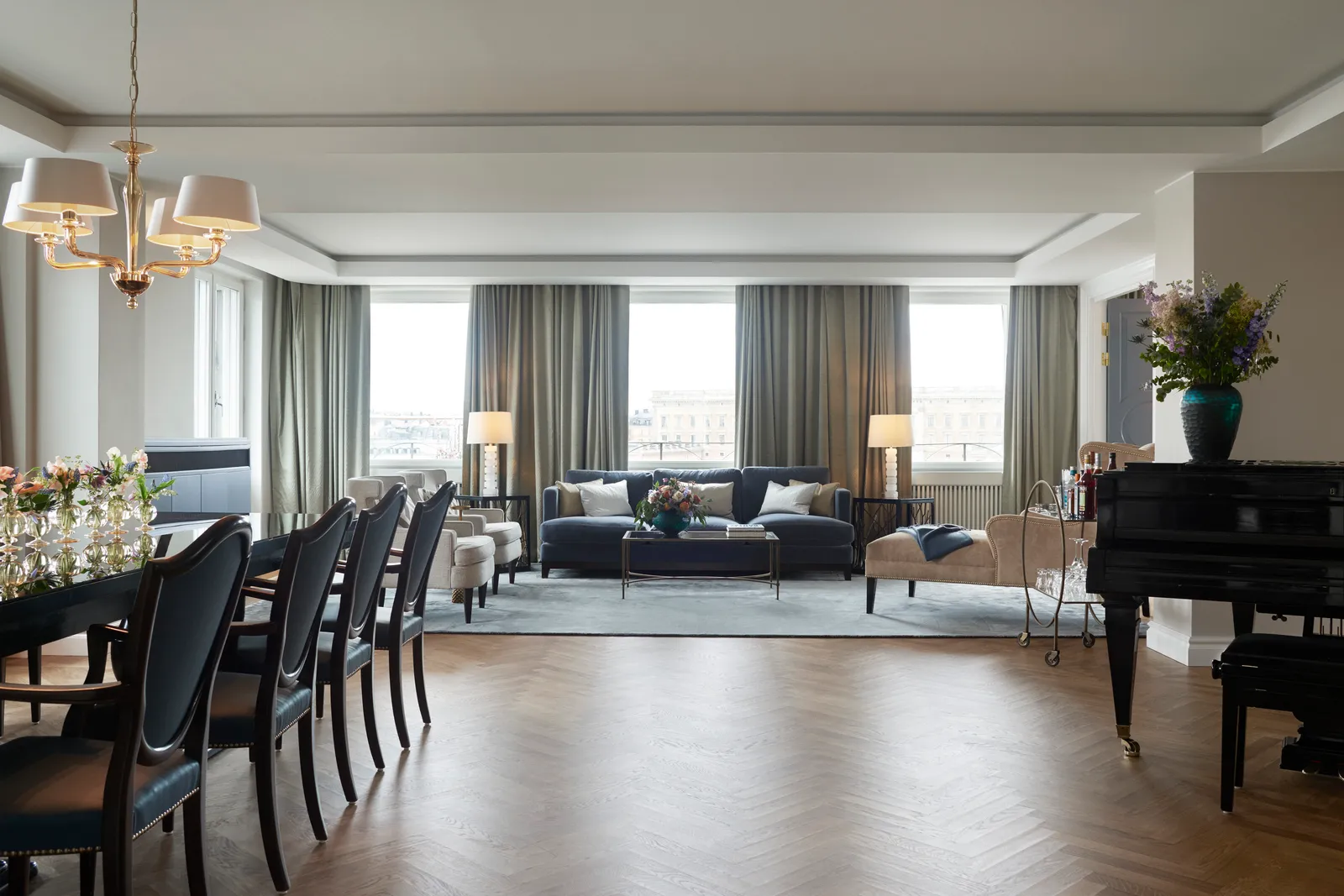 The Princess Lilian Suite living room: the long dining table, the grand piano, blue sofas and a row of windows onto the city.