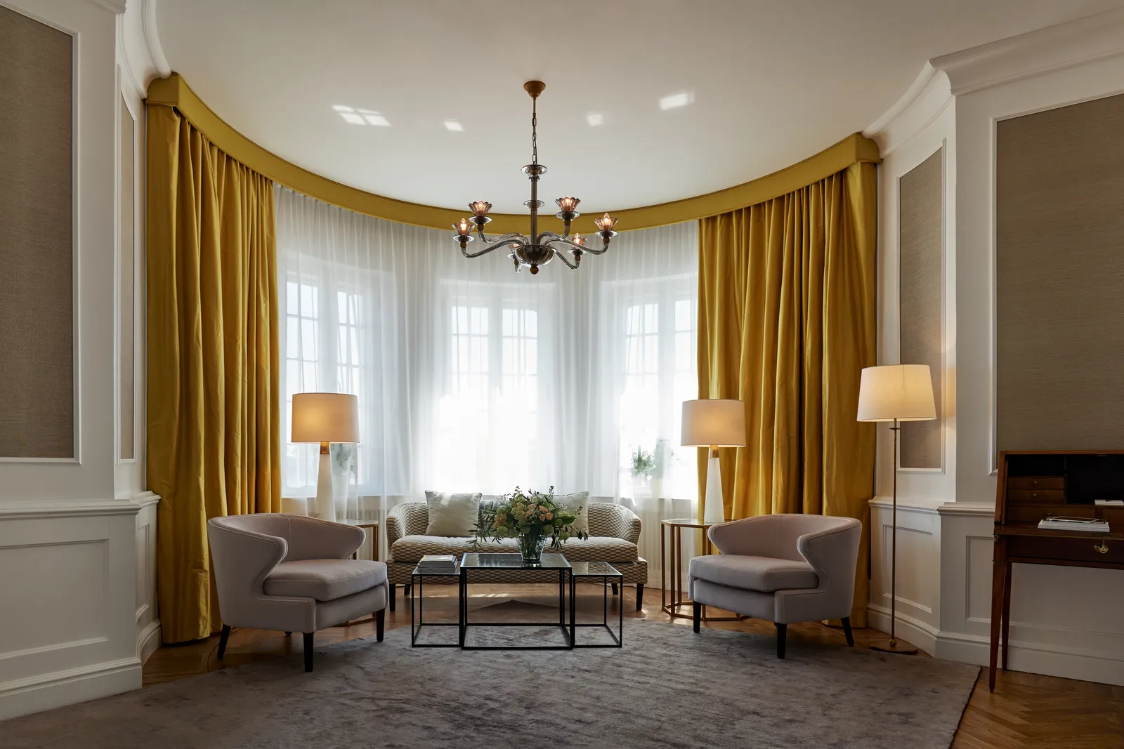 The Royal Suite sitting room: the bay window with golden-yellow curtains, 2 armchairs and a sofa around a low table, under a brass chandelier.
