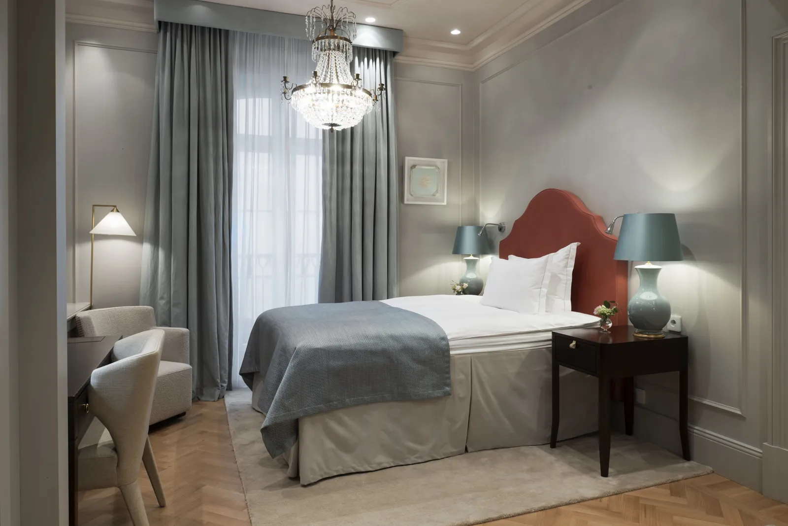 A Single Room: the bed with a brick-red headboard, grey curtains at the tall window, a small armchair and a crystal chandelier.