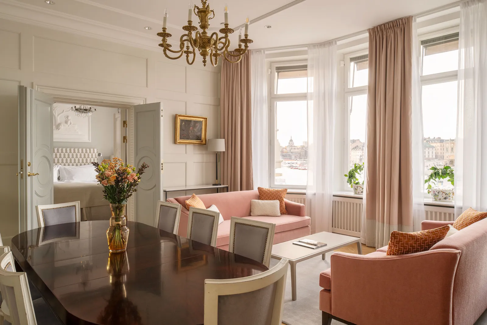 A suite parlour: the mahogany dining table, 2 pink sofas, blush curtains and 3 windows onto the water, the bedroom through the door.