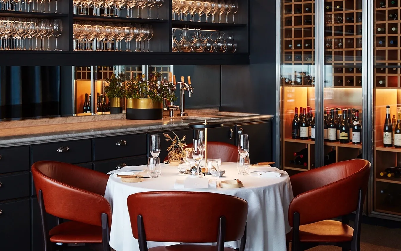 A round table laid at Seafood Gastro, with tan leather chairs, before the counter and the wall of wine.