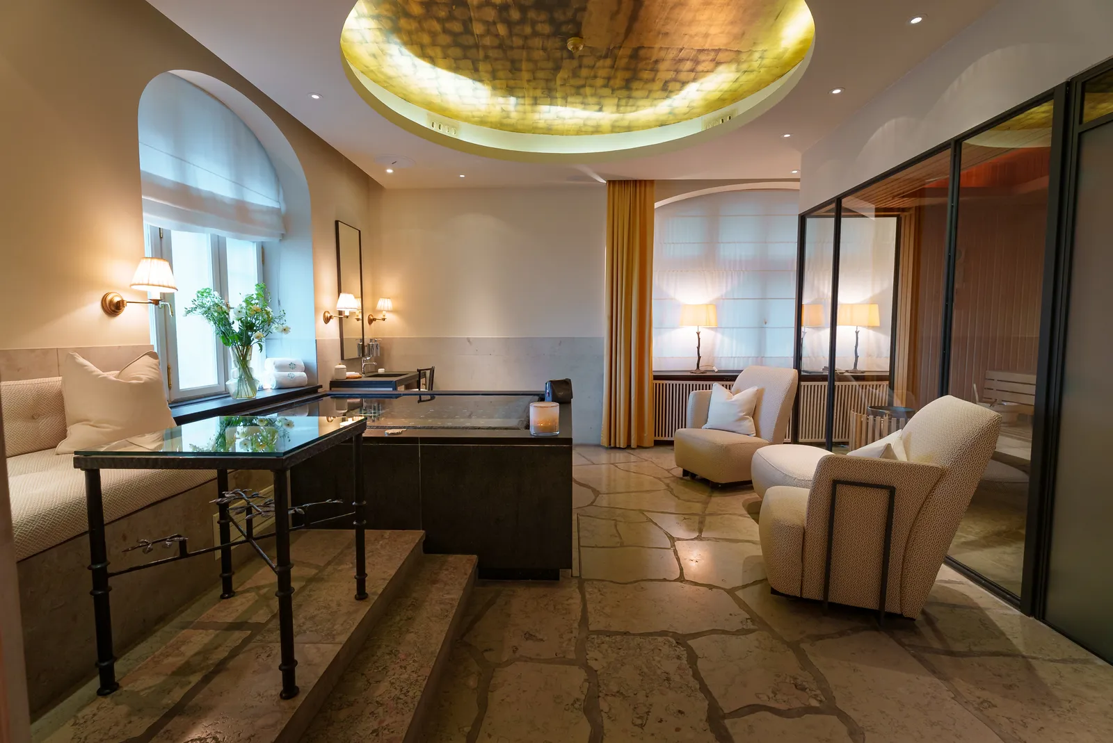 A spa suite: the dark stone pool at its centre, pale armchairs, an oval gilded ceiling and a glass wall.
