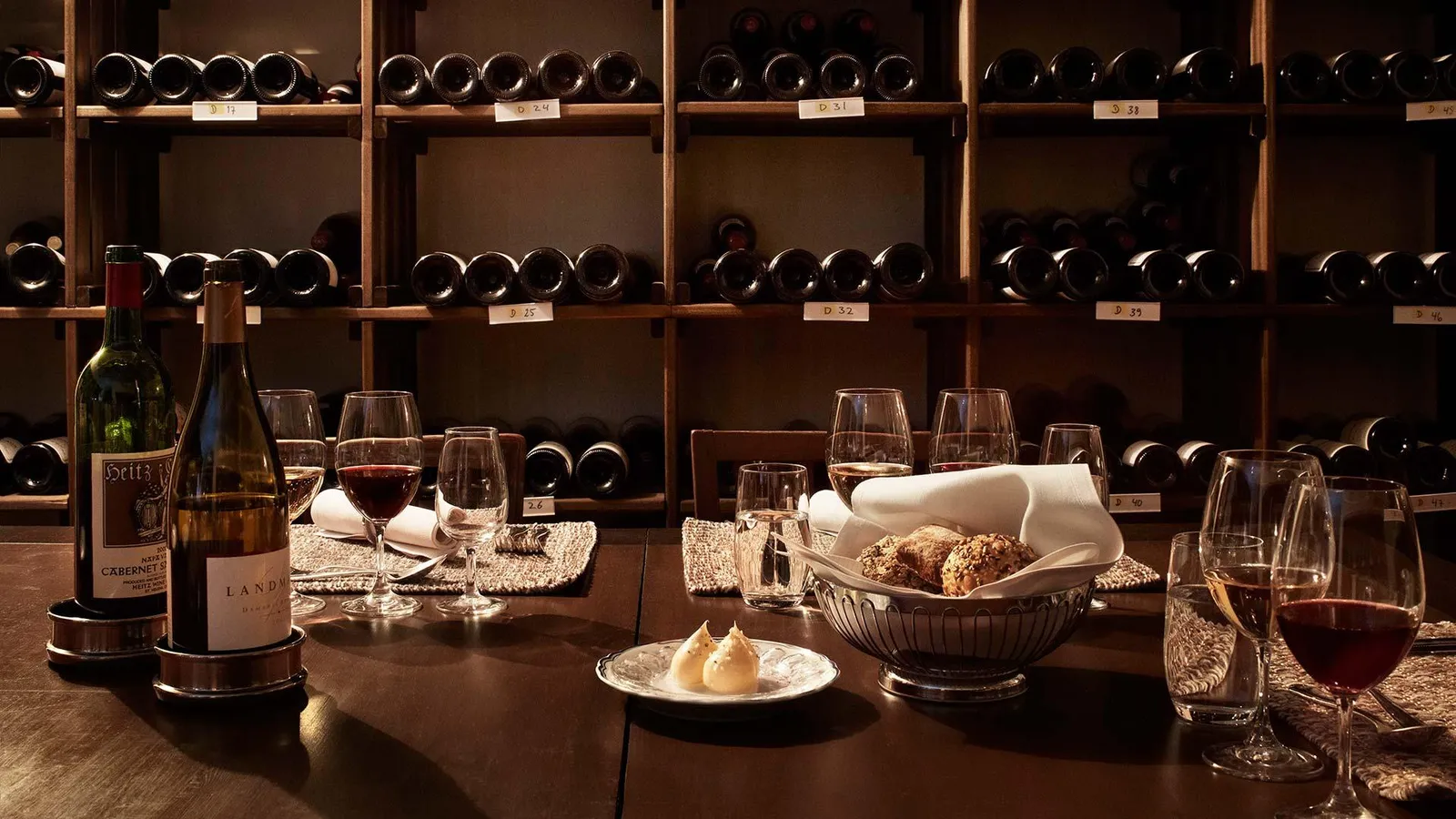The wine cellar: a table with glasses of red, a basket of bread and 2 bottles, before wooden racks full of bottles lying on their sides.