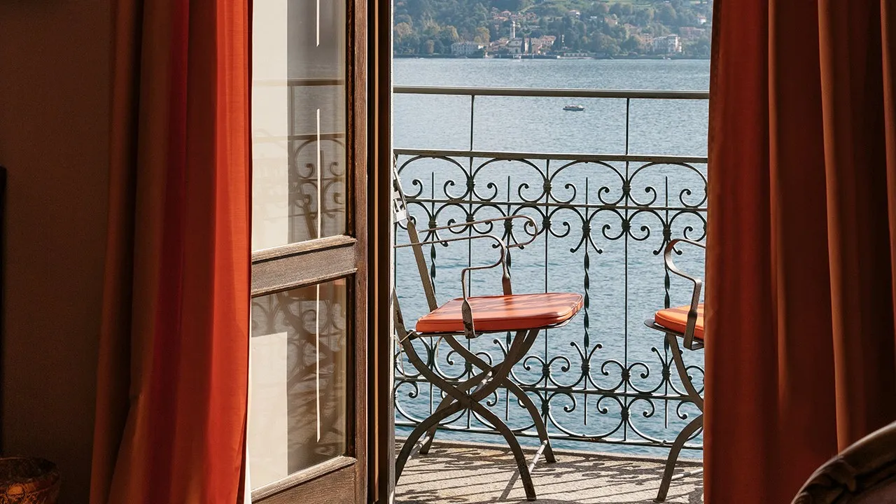 A Lake View Prestige Room’s French window open onto the balcony, with 2 iron chairs with orange cushions and the lake beyond